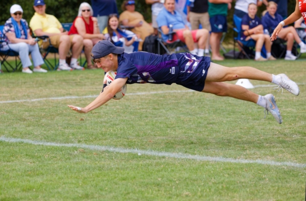 A Scotland player in a cap dives horizontally through the air with the ball, with spectators watching from camping chairs behind