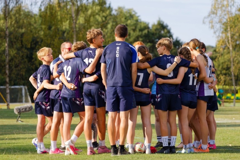 A Scotland team in navy and purple kit stand in a tight huddle with arms around each other, with a coach in a SCO shirt facing them