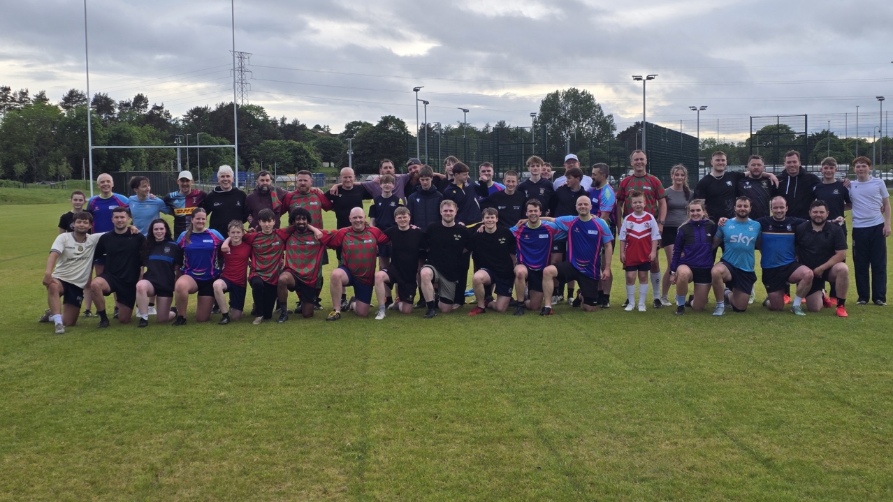 A large group of Dundee Touch players of all ages in mixed kit, some standing and some kneeling, on a grass pitch under a cloudy sky
