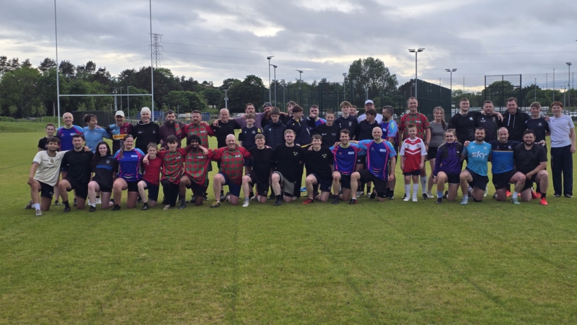 A large group of Dundee Touch players of all ages in mixed kit, some standing and some kneeling, on a grass pitch under a cloudy sky