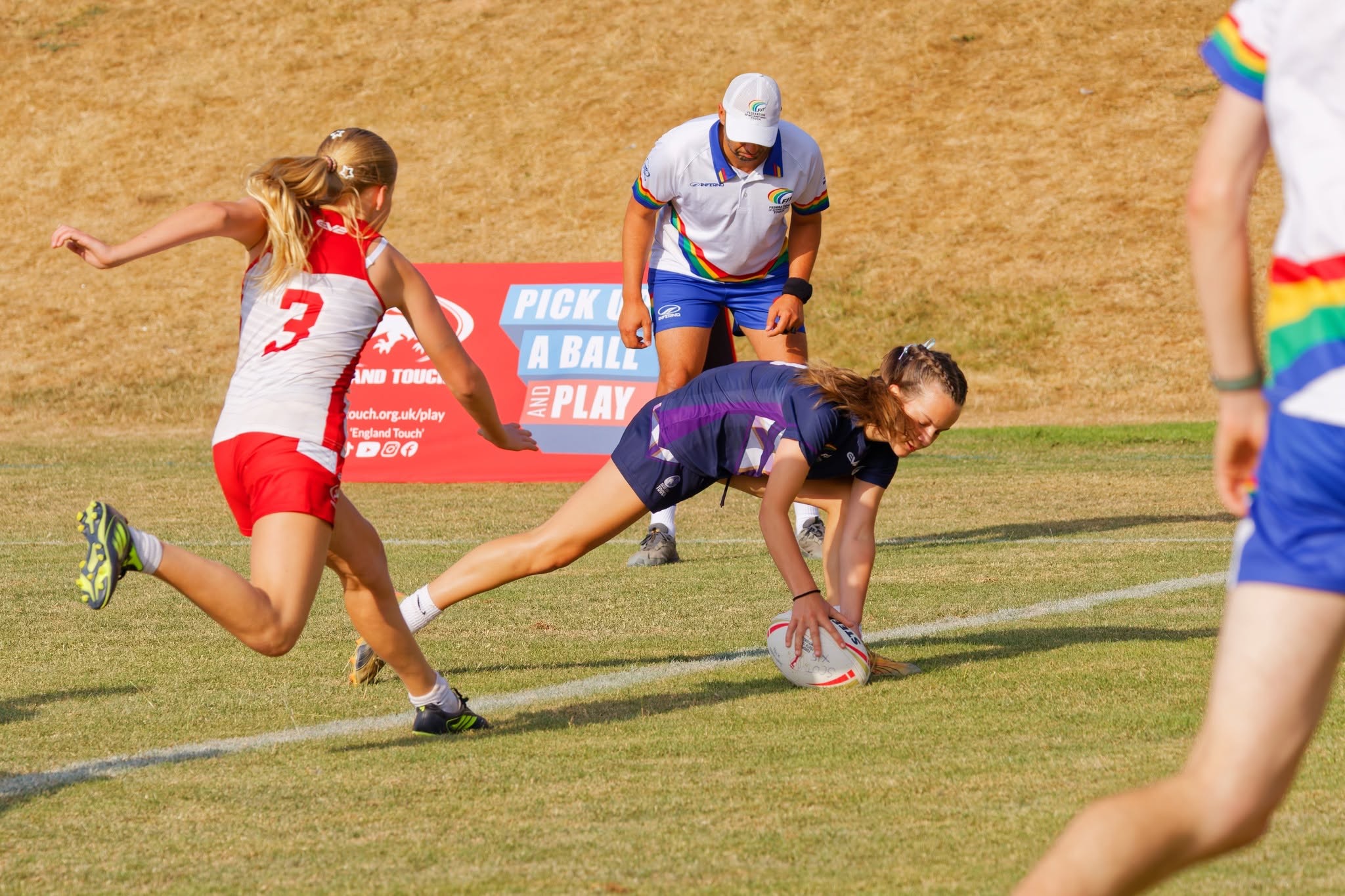 Player in navy kit reaching out to ground the ball over the line while an opponent chases and a referee looks on